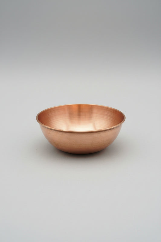 A round copper-colored metal bowl with a brushed metallic finish, placed on a light wood tabletop inside a store.