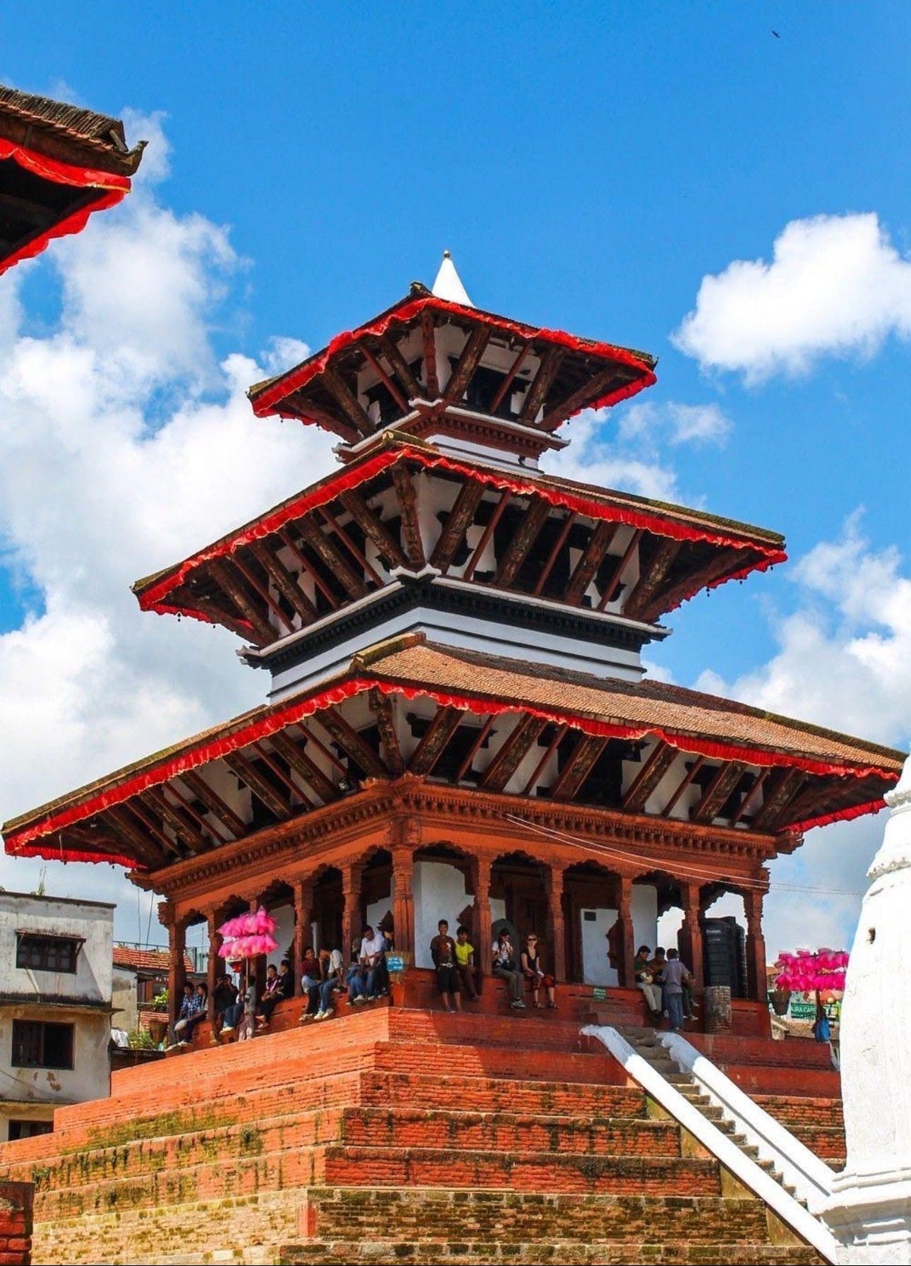A traditional multi-tiered wooden temple with red-painted brick walls and red roof trim, set against a bright blue sky with white clouds. Small groups of people are visible walking along the temple's staircase and under its covered walkway.