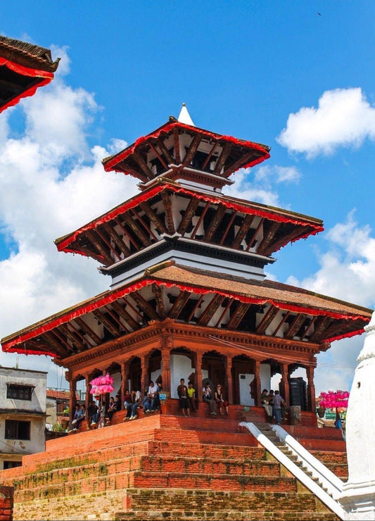A traditional multi-tiered wooden temple with red-painted brick walls and red roof trim, set against a bright blue sky with white clouds. Small groups of people are visible walking along the temple's staircase and under its covered walkway.