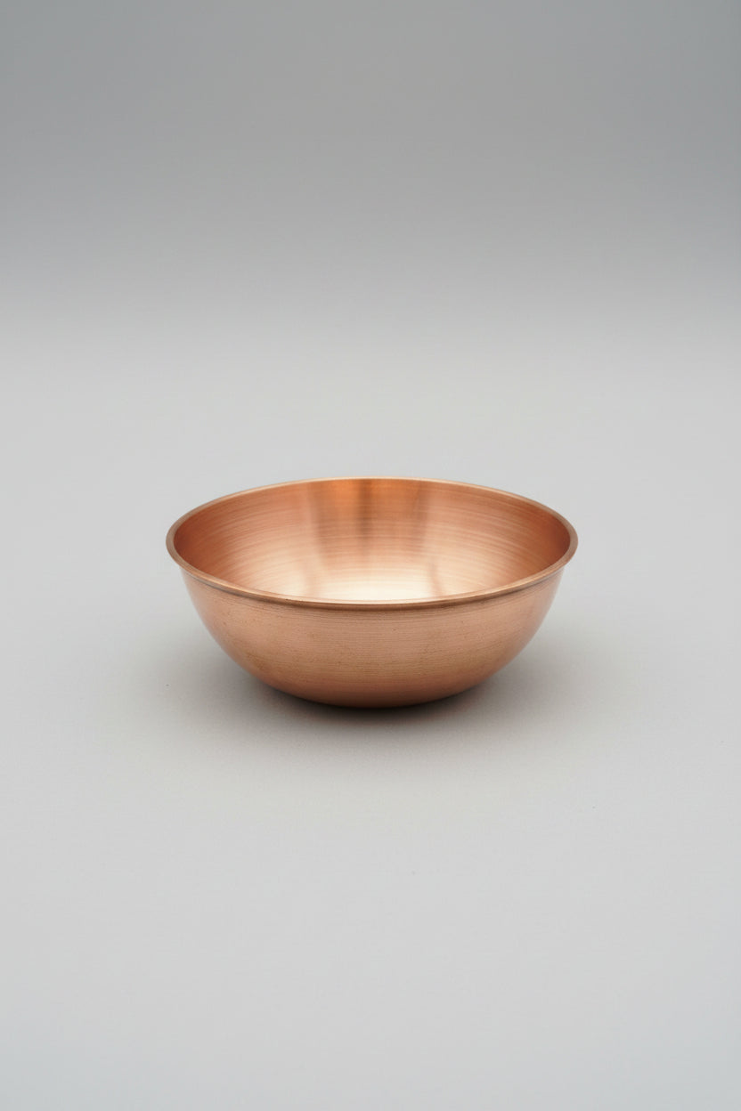 A round copper-colored metal bowl with a brushed metallic finish, placed on a light wood tabletop inside a store.