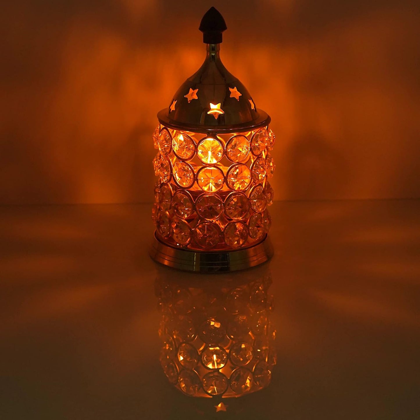 A decorative table lamp with a metallic top and base, covered in faceted circular crystal-like beads, lit from within to cast a warm orange glow and shadow on the background.