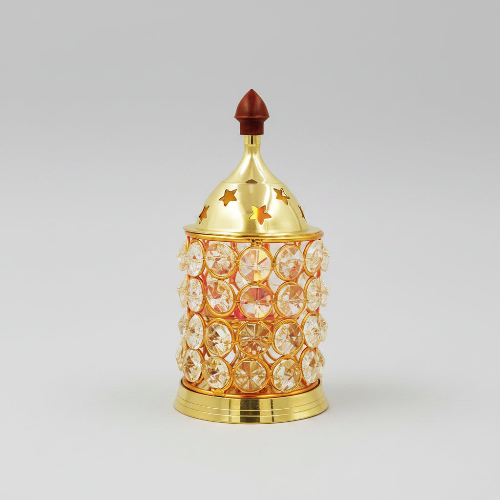 A decorative gold-toned metal incense burner with clear rhinestone-like circular embellishments and star cutouts on the lid, styled on a tabletop with stacked stones, green leaves, a pink flower, and a rolled bamboo mat against a softly blurred bamboo background.