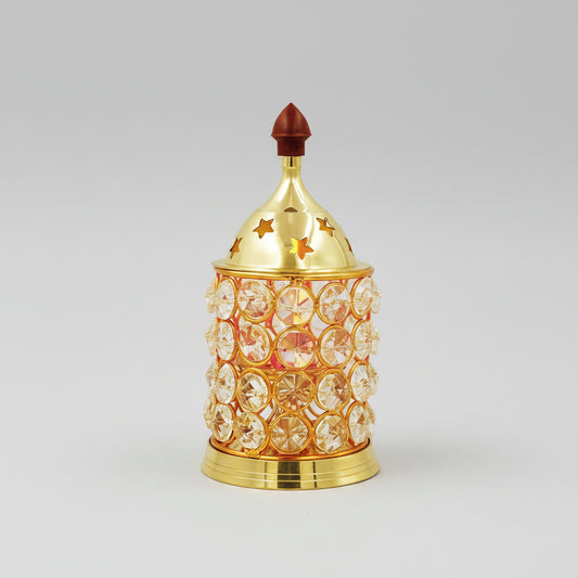 A decorative gold-toned metal incense burner with clear rhinestone-like circular embellishments and star cutouts on the lid, styled on a tabletop with stacked stones, green leaves, a pink flower, and a rolled bamboo mat against a softly blurred bamboo background.